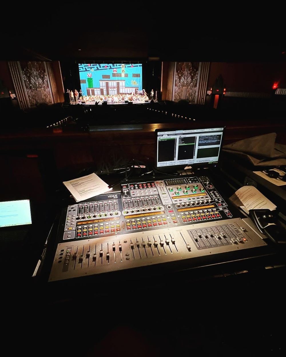 Front of house at a theatre show, mixing console in the foreground and the stage lit in the distance